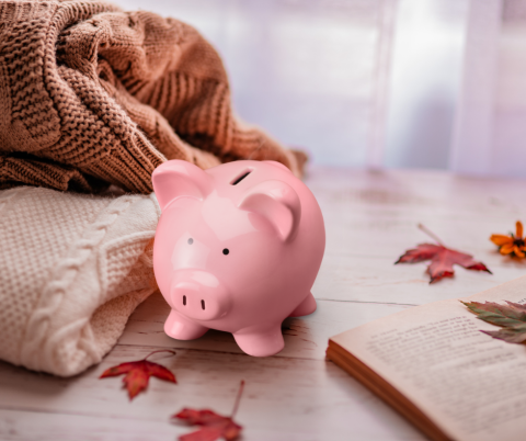 Table with folded sweaters, open book, fallen leaves, and a piggy bank